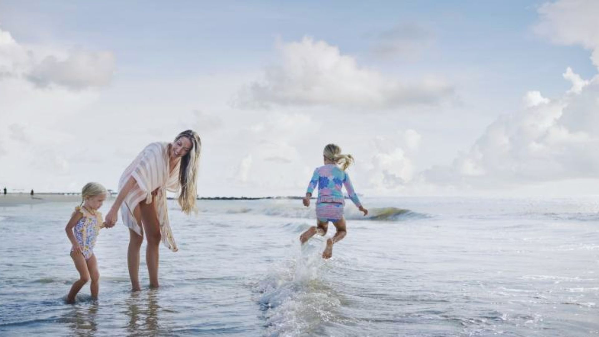 Parent and children play in shallow ocean water on Tybee Island beach under bright cloudy skies.