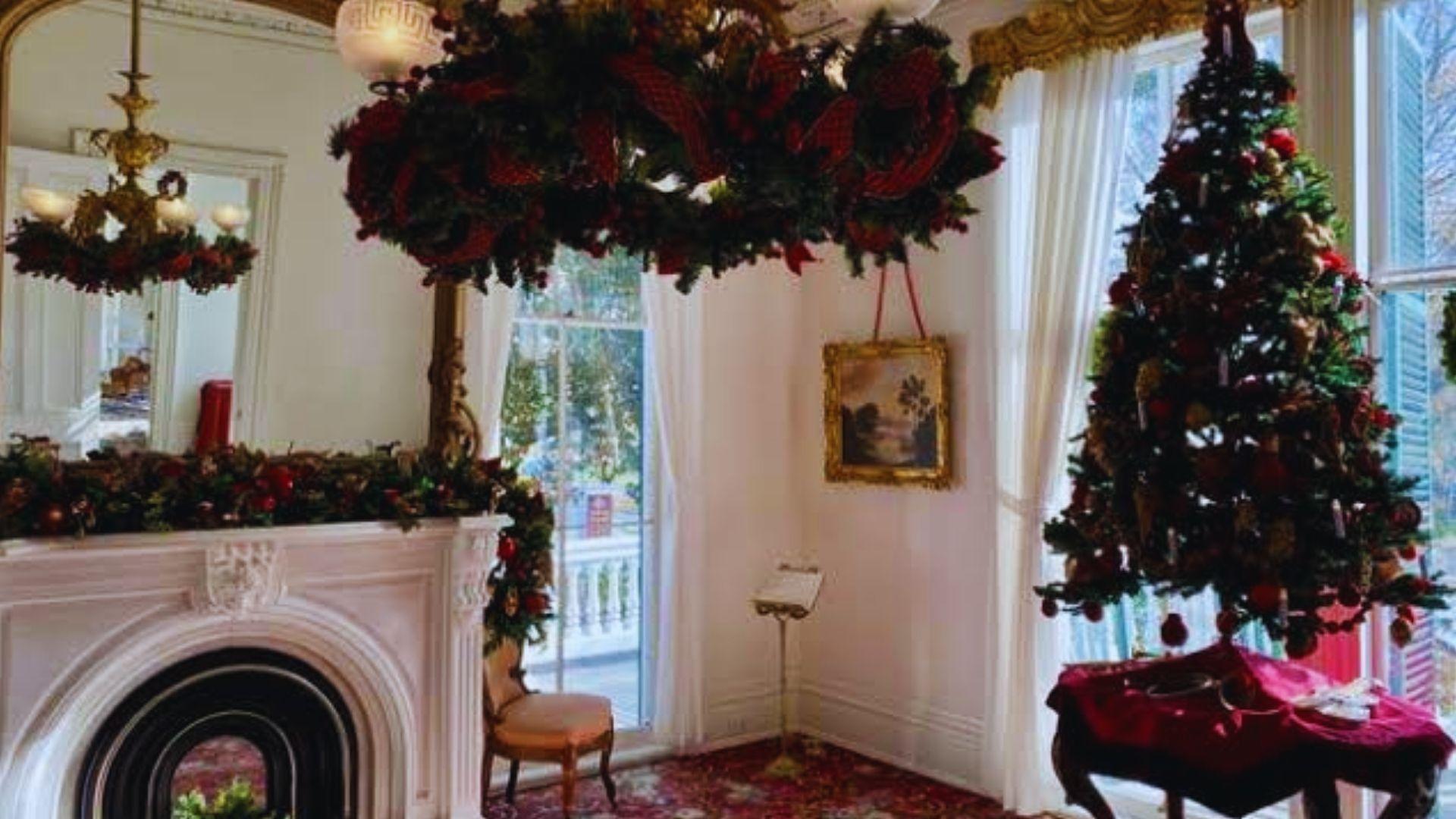 Victorian parlor decorated for Christmas with ornate fireplace garland, chandelier wreath, Christmas tree, mirror, sunlit windows