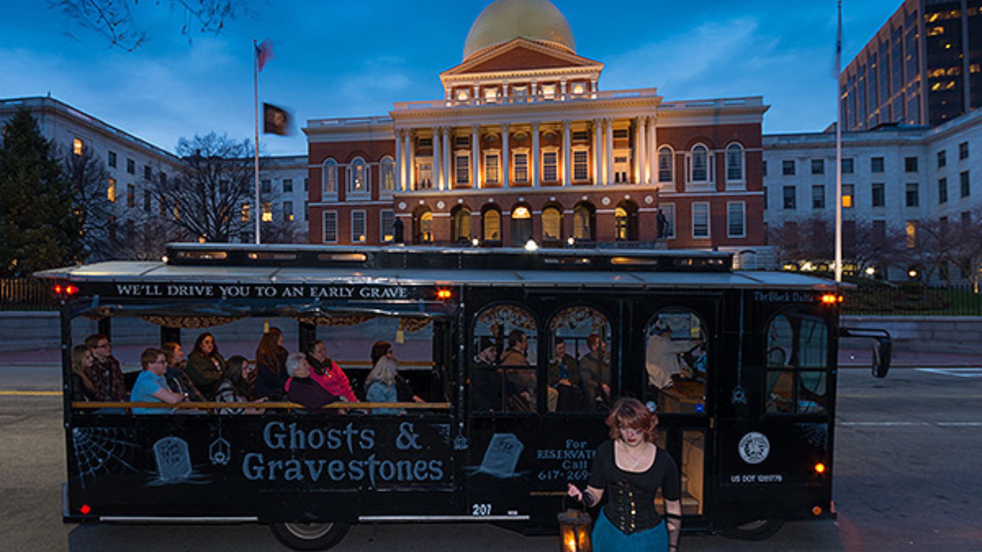 . Black Ghosts & Gravestones trolley bus with tourists at dusk in front of the illuminated Massachusetts State House with gold dome.
