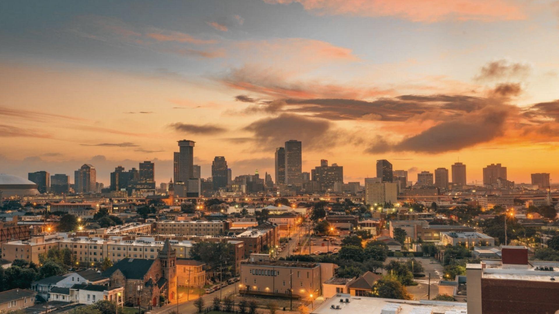 New Orleans downtown skyline at sunset with Caesars Superdome, skyscrapers, streetlights glowing, orange clouds, residential neighborhoods below