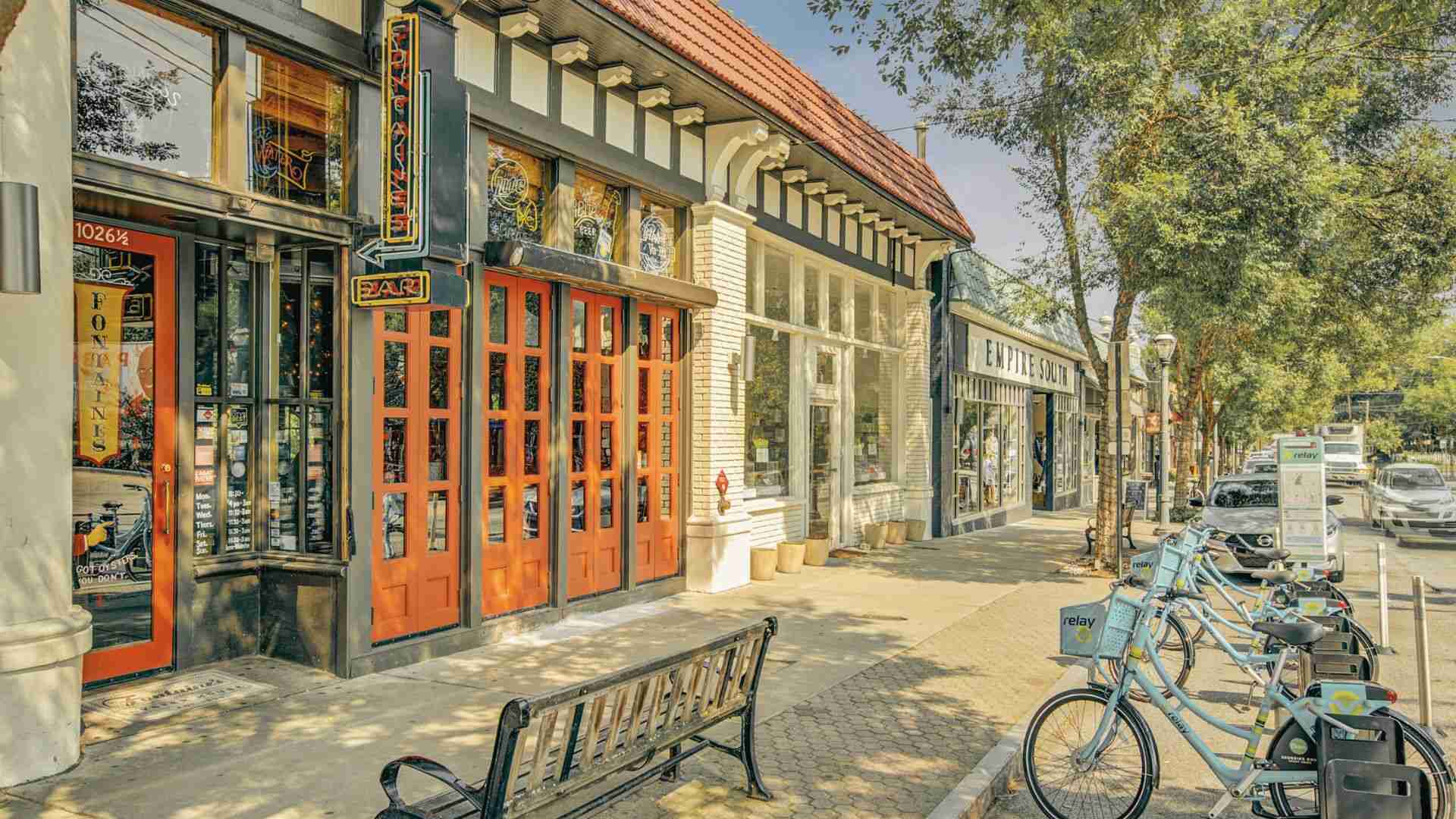 Virginia Highland streetscape with storefronts, colorful doors, shaded sidewalks, and shared bicycles parked outside shops.