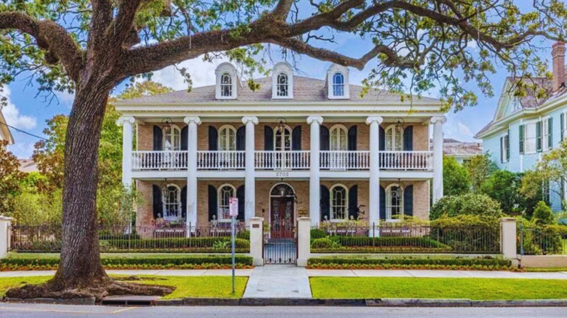 Two-story brick Greek Revival mansion with white columns, black shutters, gated yard, large oak tree, address 2702