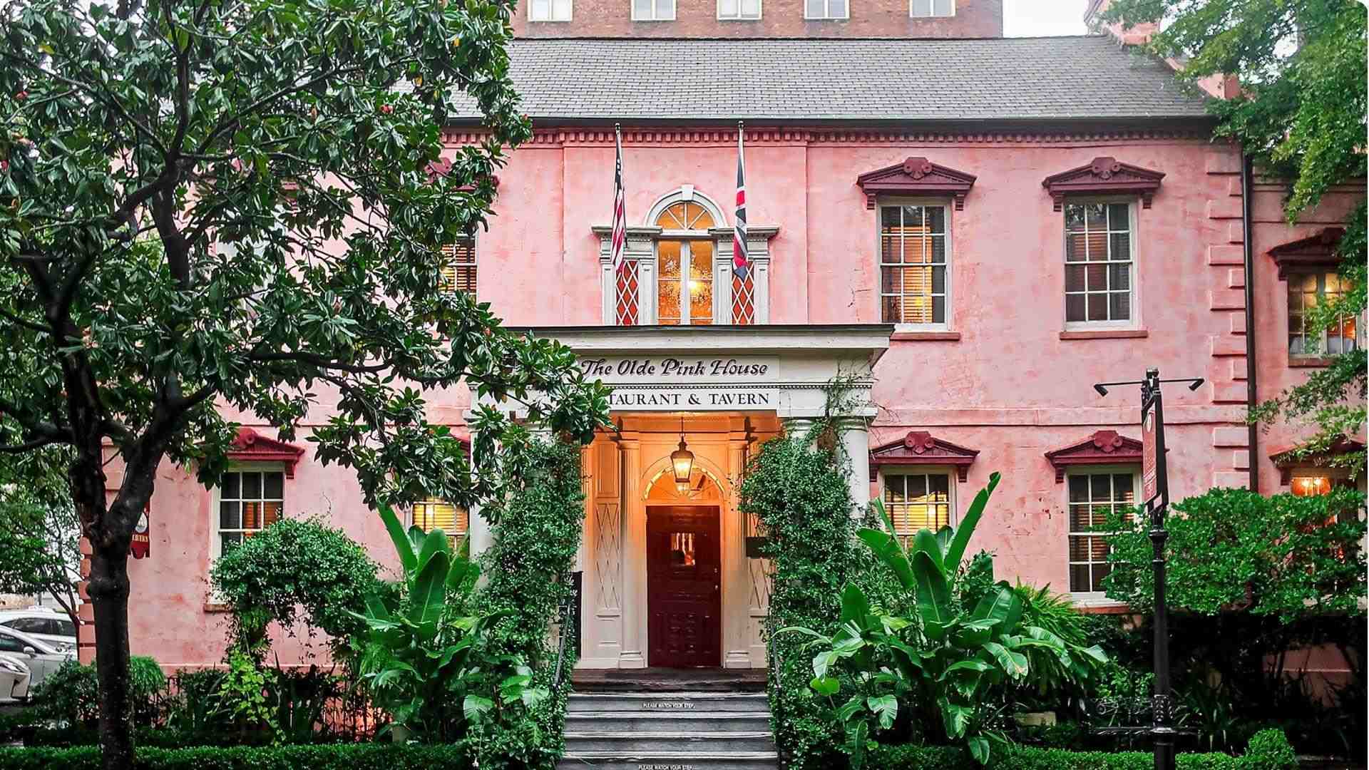 A historic pink stucco Georgian mansion with white trim, lantern-lit entrance, and greenery in Savannah, GA.