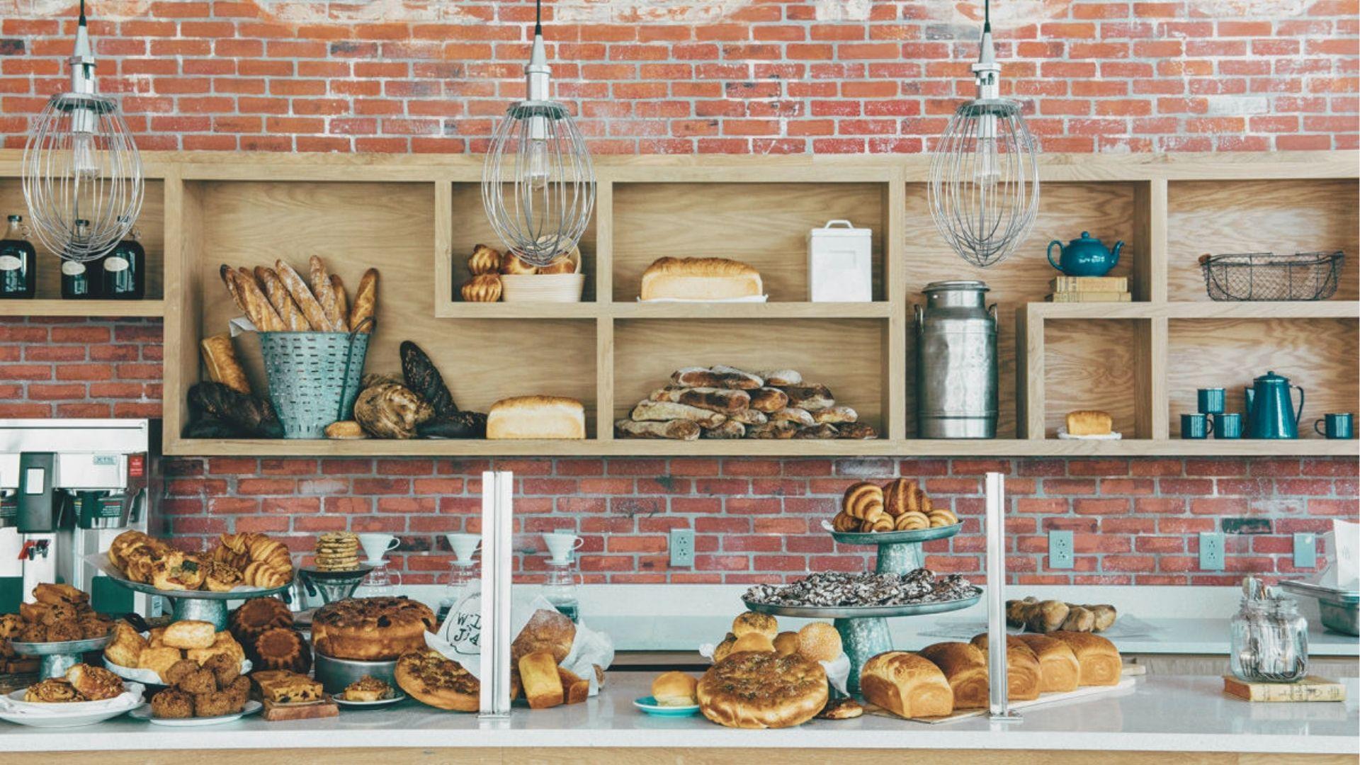 Rustic bakery display with artisan breads, croissants, and pastries stacked on shelves against a brick wall.