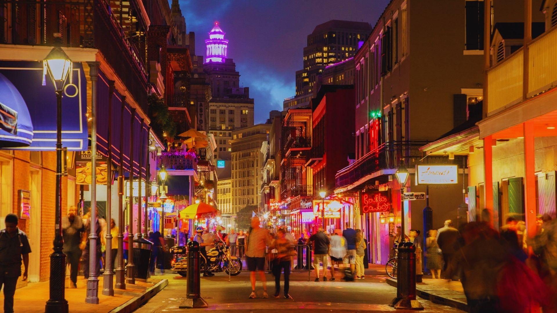 Bourbon Street at night with neon signs, iron balconies, crowds walking, gas lamps glowing, purple-lit tower visible downtown