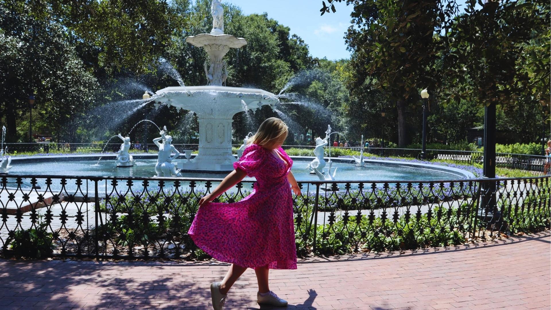 Woman in pink dress twirling by Forsyth Park fountain on a sunny day in Savannah.