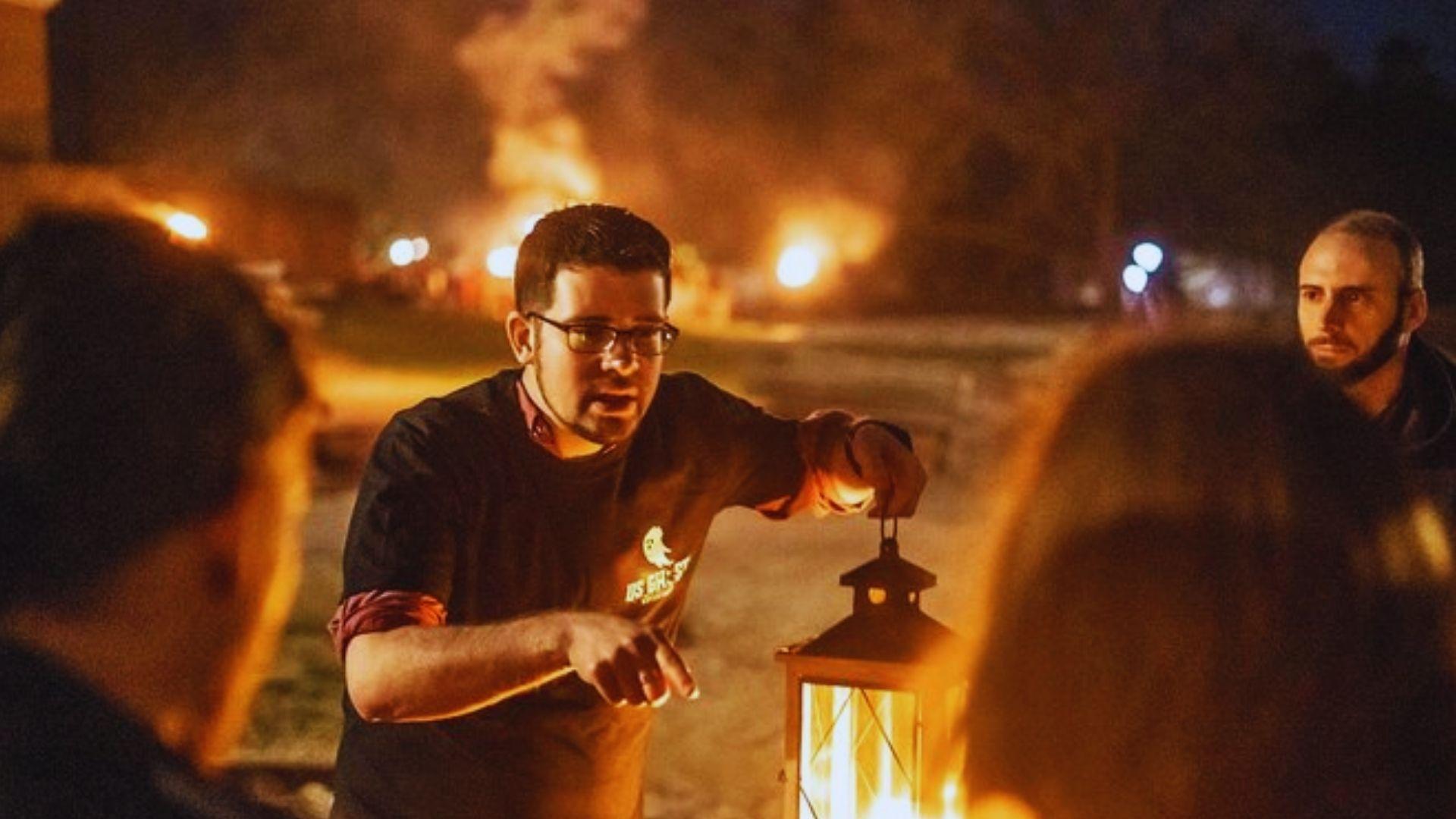 Night ghost tour guide with lantern gestures to group while bonfires blaze on levee, orange light, crowd