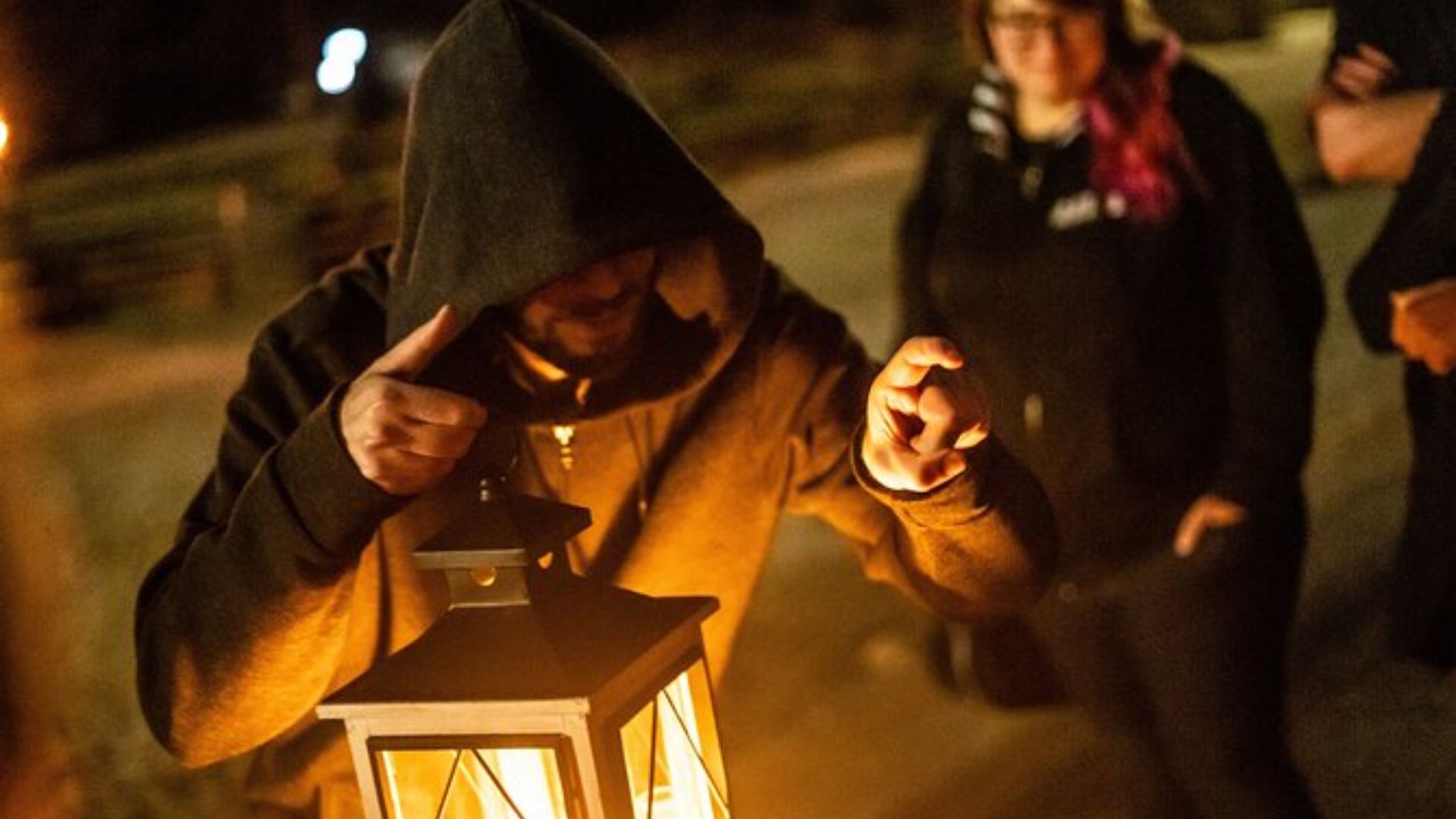 Hooded man holding a glowing lantern at night, pointing forward while two people stand blurred in background.