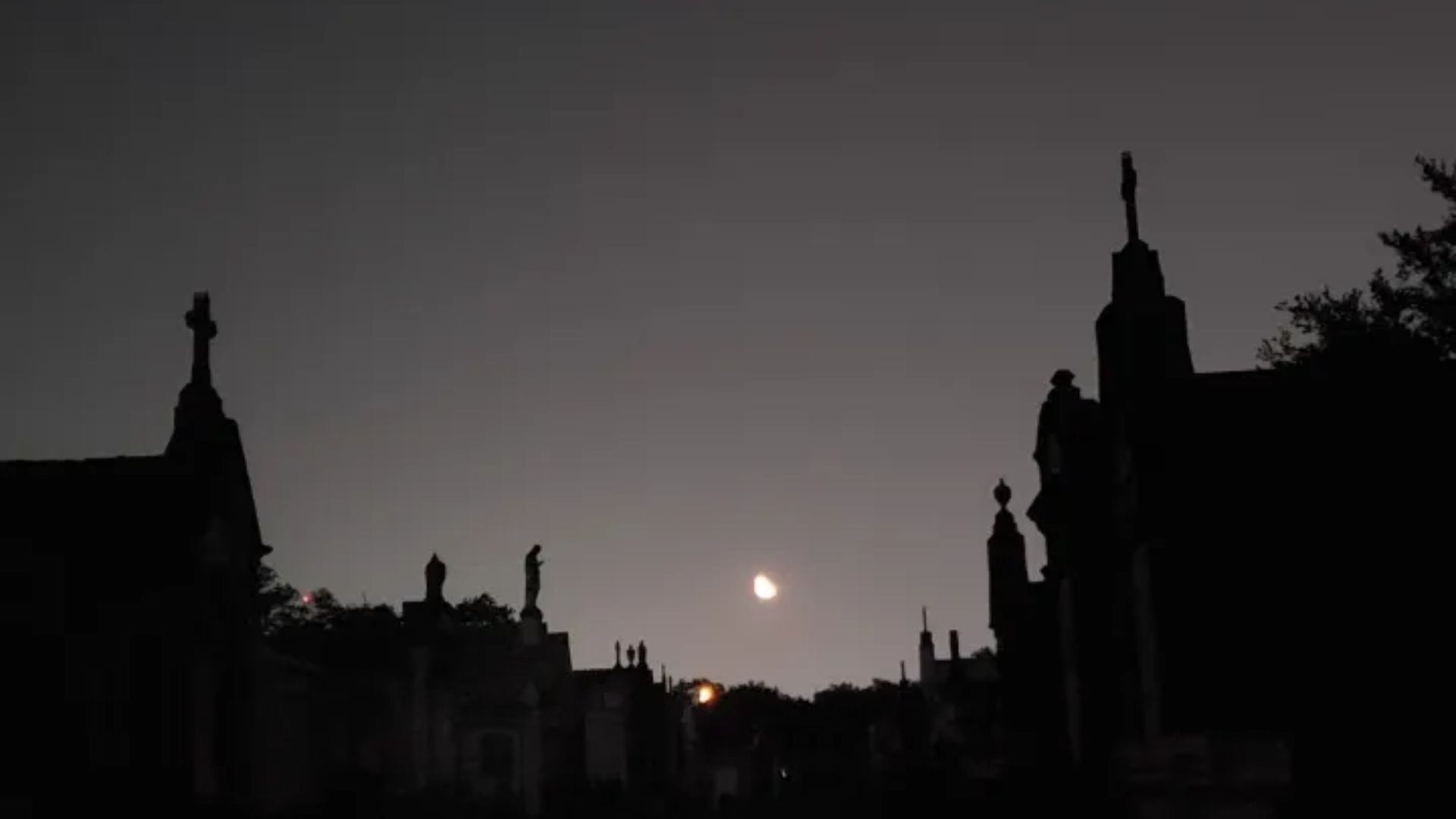 Silhouetted cemetery mausoleums and statues under a dark twilight sky with a bright moon glowing between tombs.