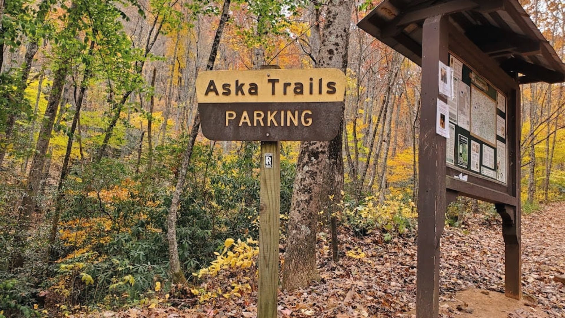 Aska Trails parking sign stands beside an information board in a forest with autumn colored trees nearby.