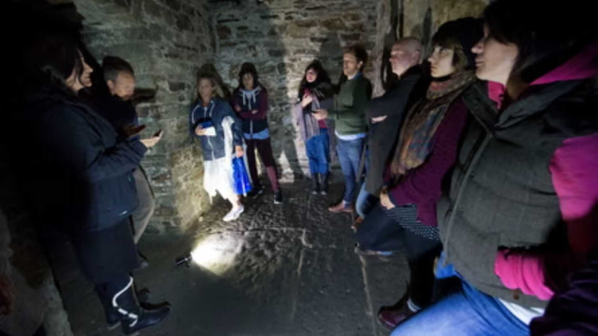 Tour group gathered in a dimly lit stone vault, listening to a guide holding a phone with a flashlight on the floor.