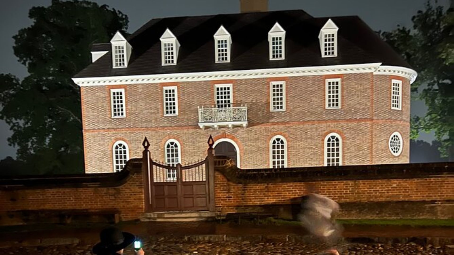 Historic brick mansion lit at night behind a wall, with a blurry ghost-like figure and a person in a hat taking a photo.