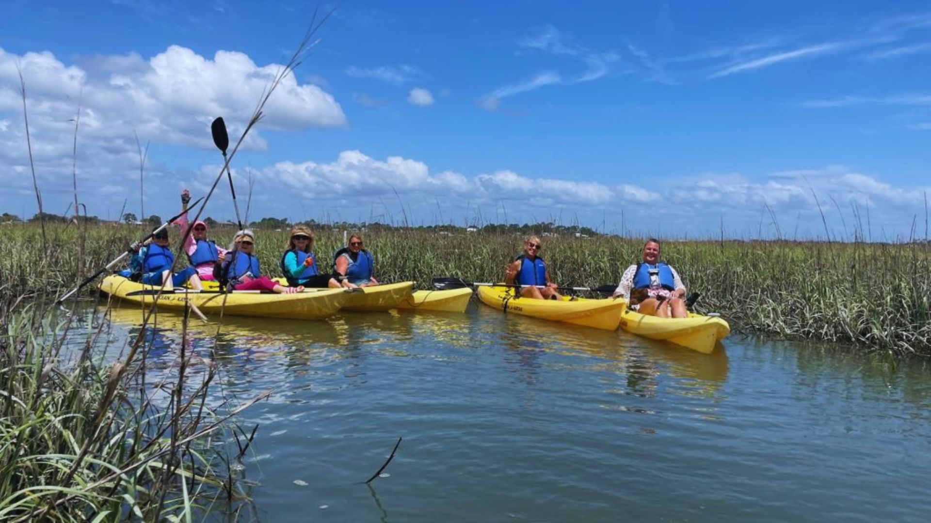 People in yellow kayaks paddle through marsh water under blue skies and white clouds together.