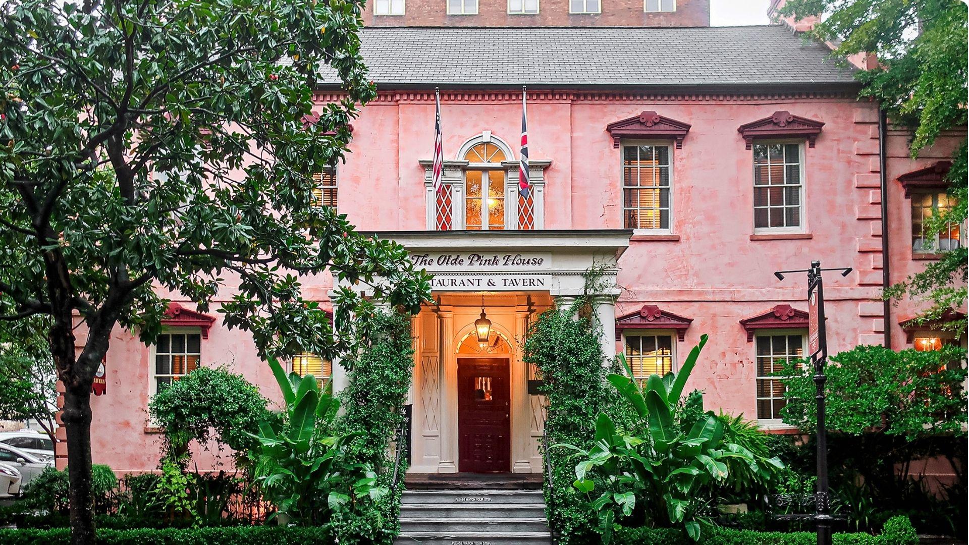 A historic pink stucco Georgian mansion with white trim, lantern-lit entrance, and lush tropical greenery in Savannah, GA.