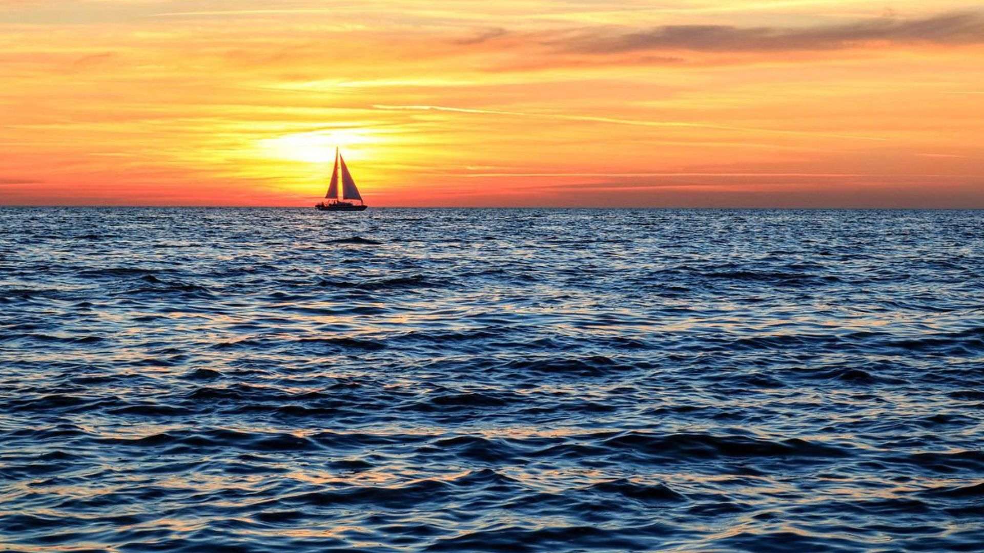 A sailboat glides on calm ocean water in front of an orange sunset.