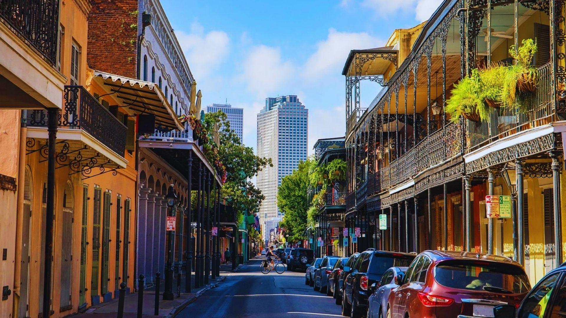 Colorful French Quarter street with iron balconies, hanging ferns, parked cars, cyclist, skyscrapers in distance, sunshine