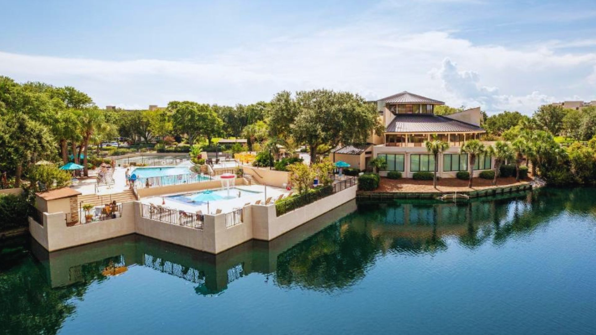 Resort building and outdoor pools beside a calm lagoon, surrounded by trees and palm plants, at Island Club of Hilton Head Island.