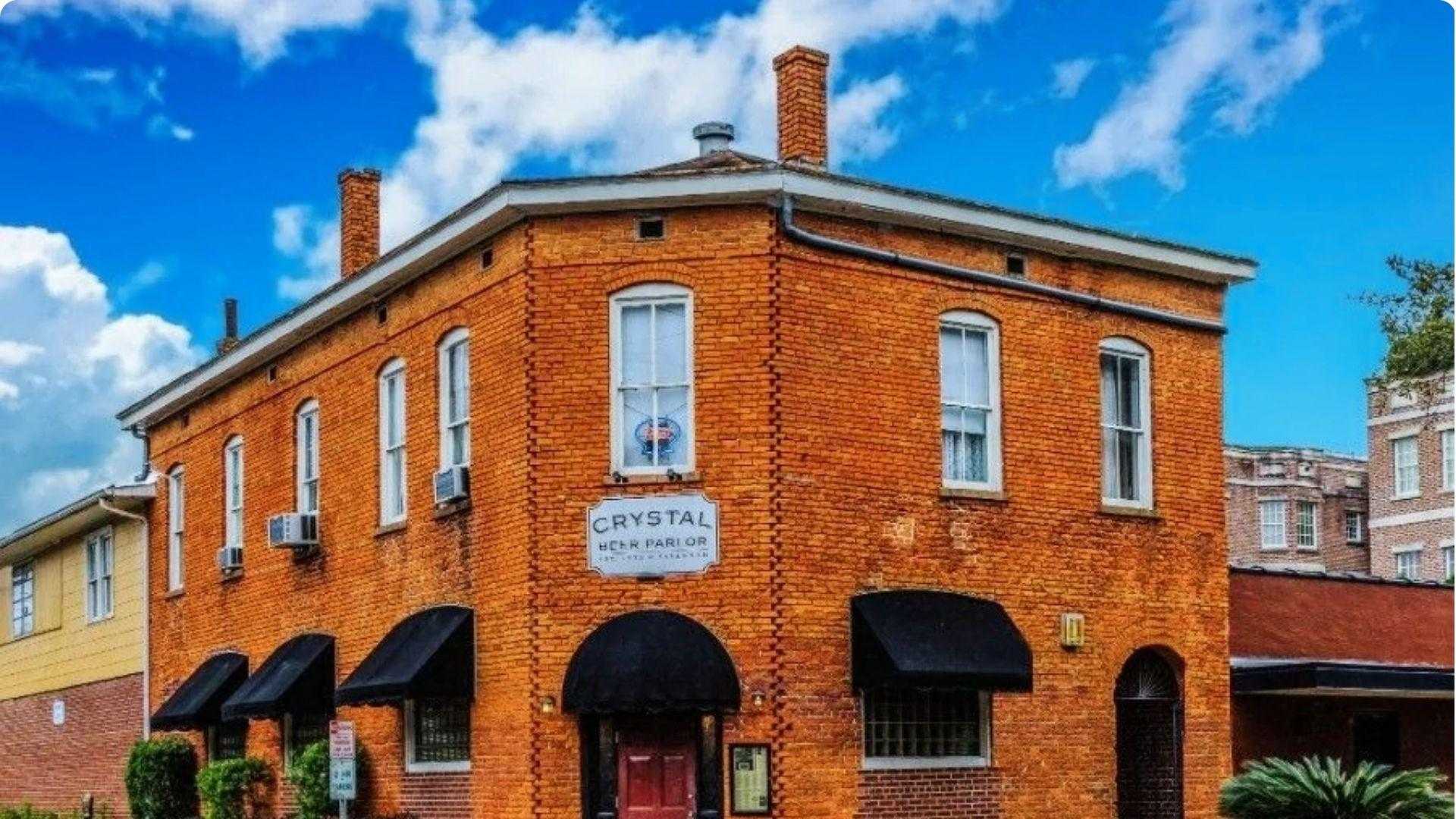 A two-story red brick corner building with black awnings and a vintage sign, one of Savannah's oldest bars.