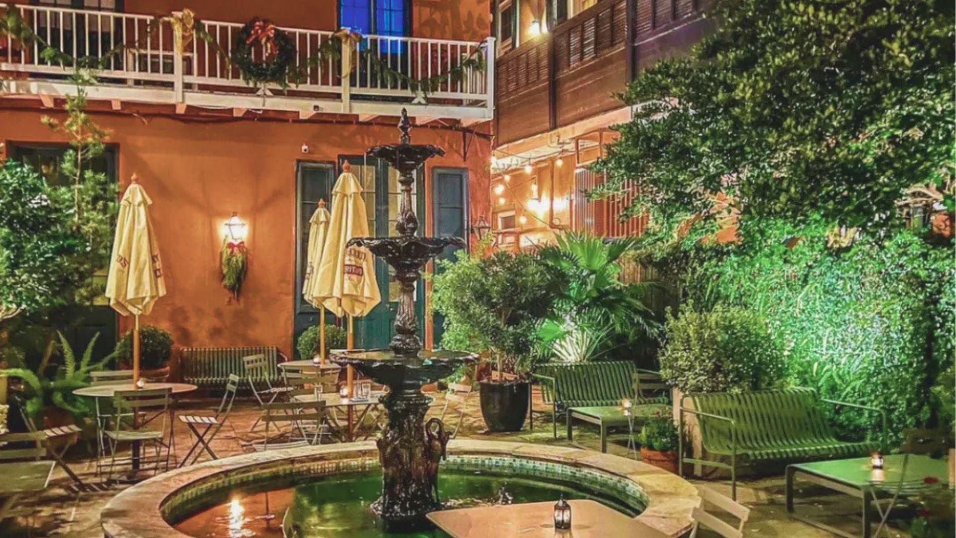 Romantic courtyard dining area at night centered on a three-tier fountain, surrounded by lush plants and string lights.