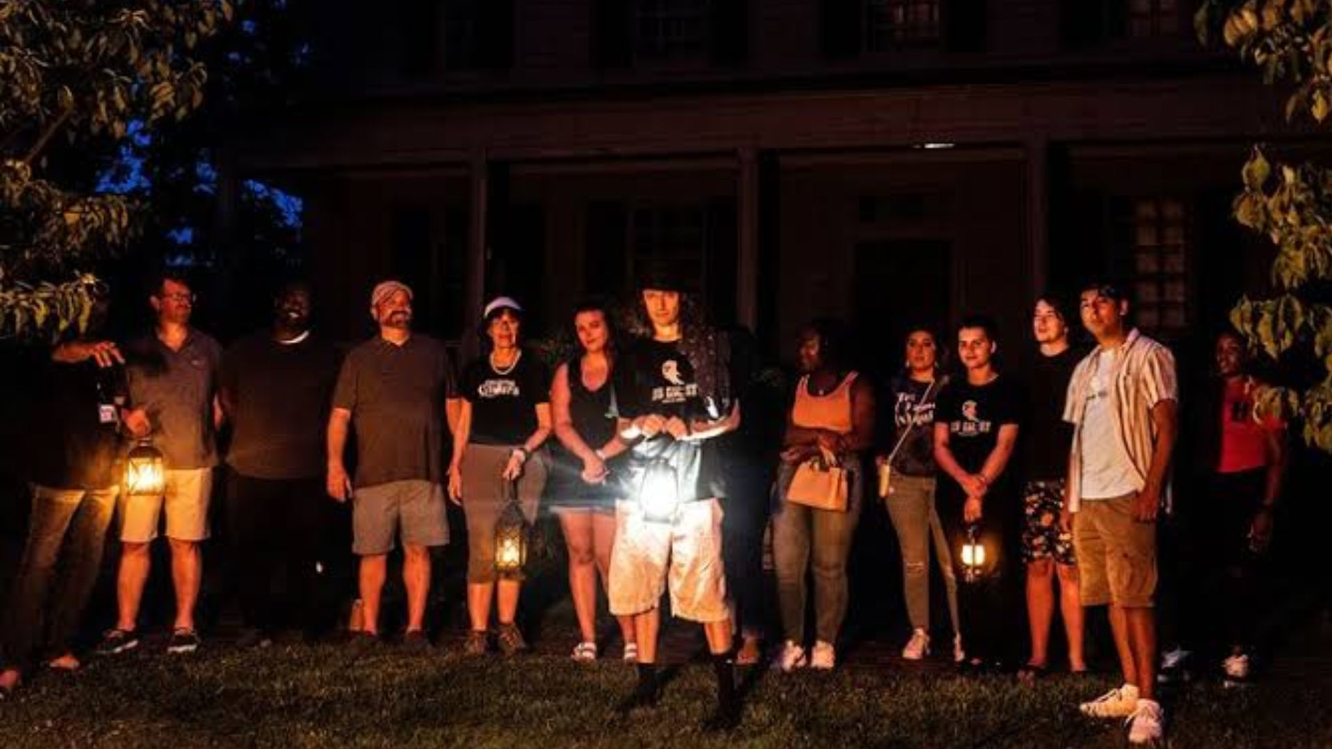 Group of people holding lanterns on a lawn at night outside a dark house, listening to a central guide.