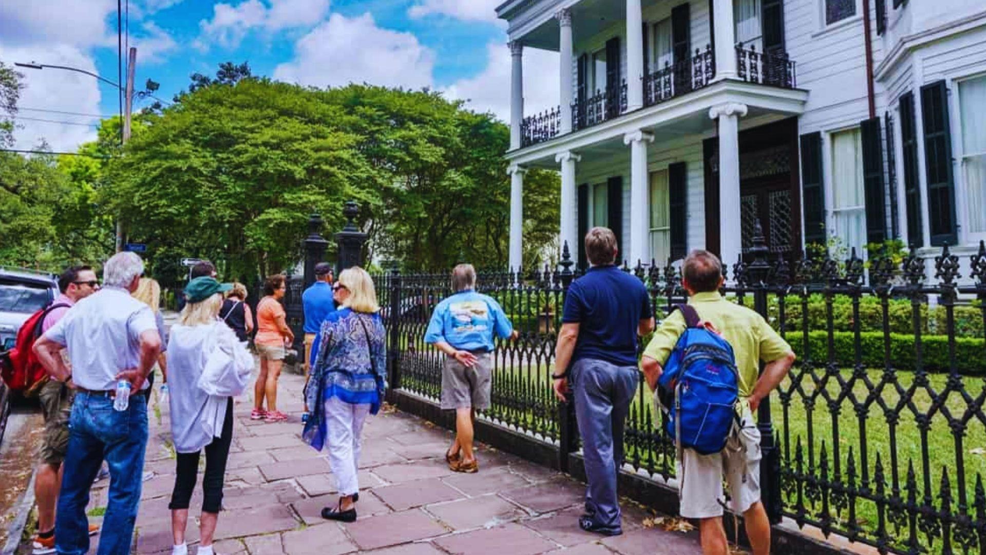 Tour group gathers on sidewalk admiring white Garden District mansion with columns, black iron fence, blue sky