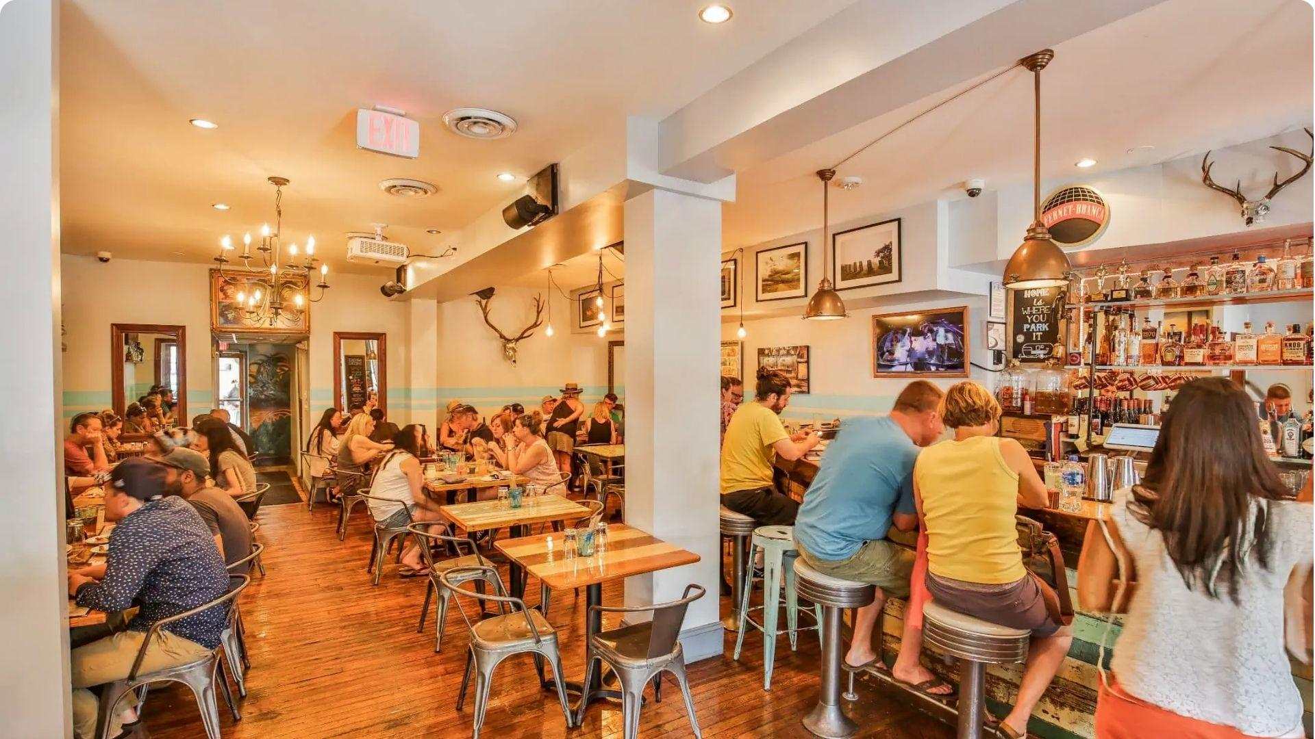 Lively, warmly lit casual restaurant interior packed with diners at wooden tables and bar stools.