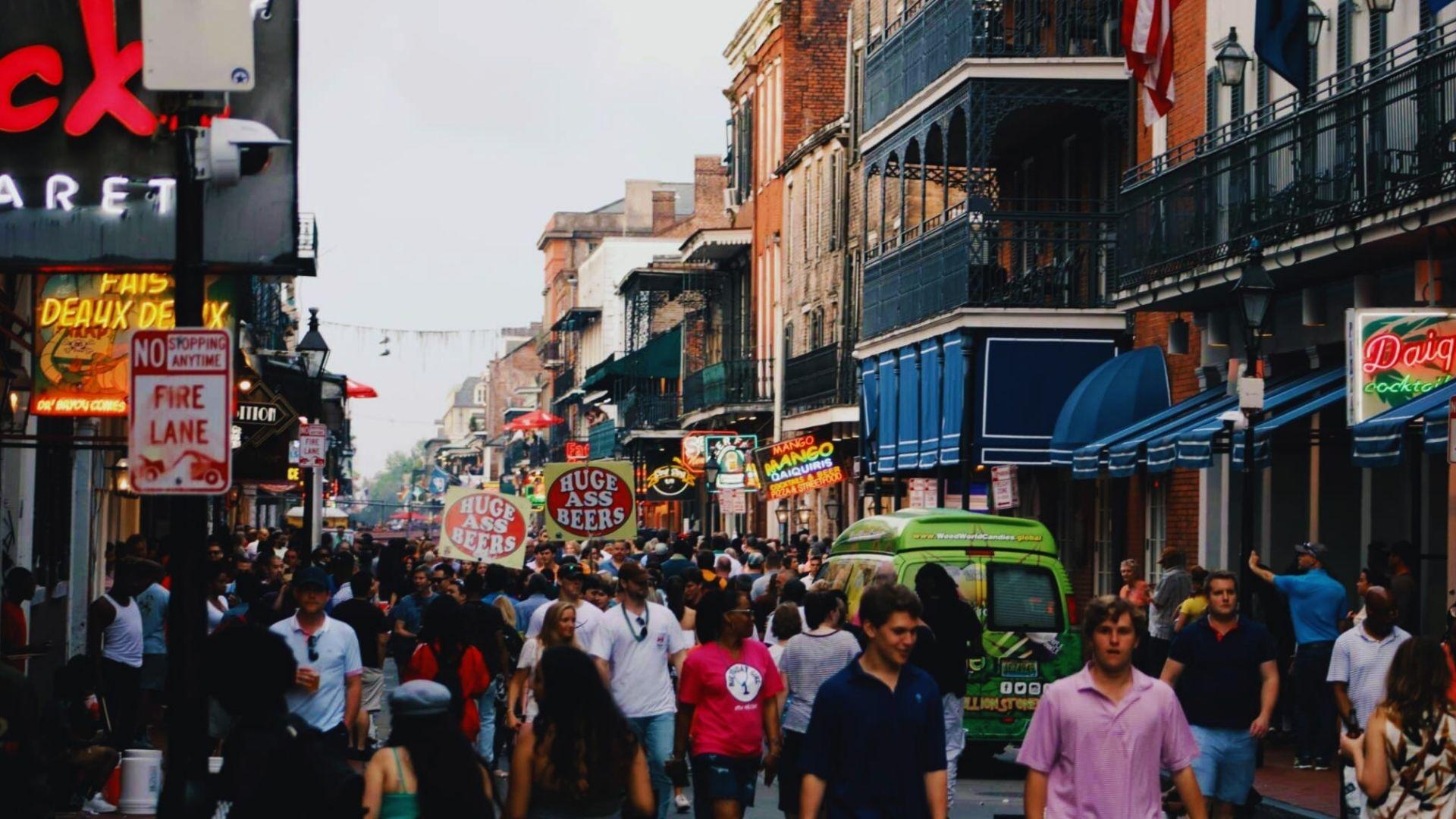 Crowded Bourbon Street with neon signs, iron balconies, "Huge Ass Beers" signs, tourists walking, daytime, French Quarter