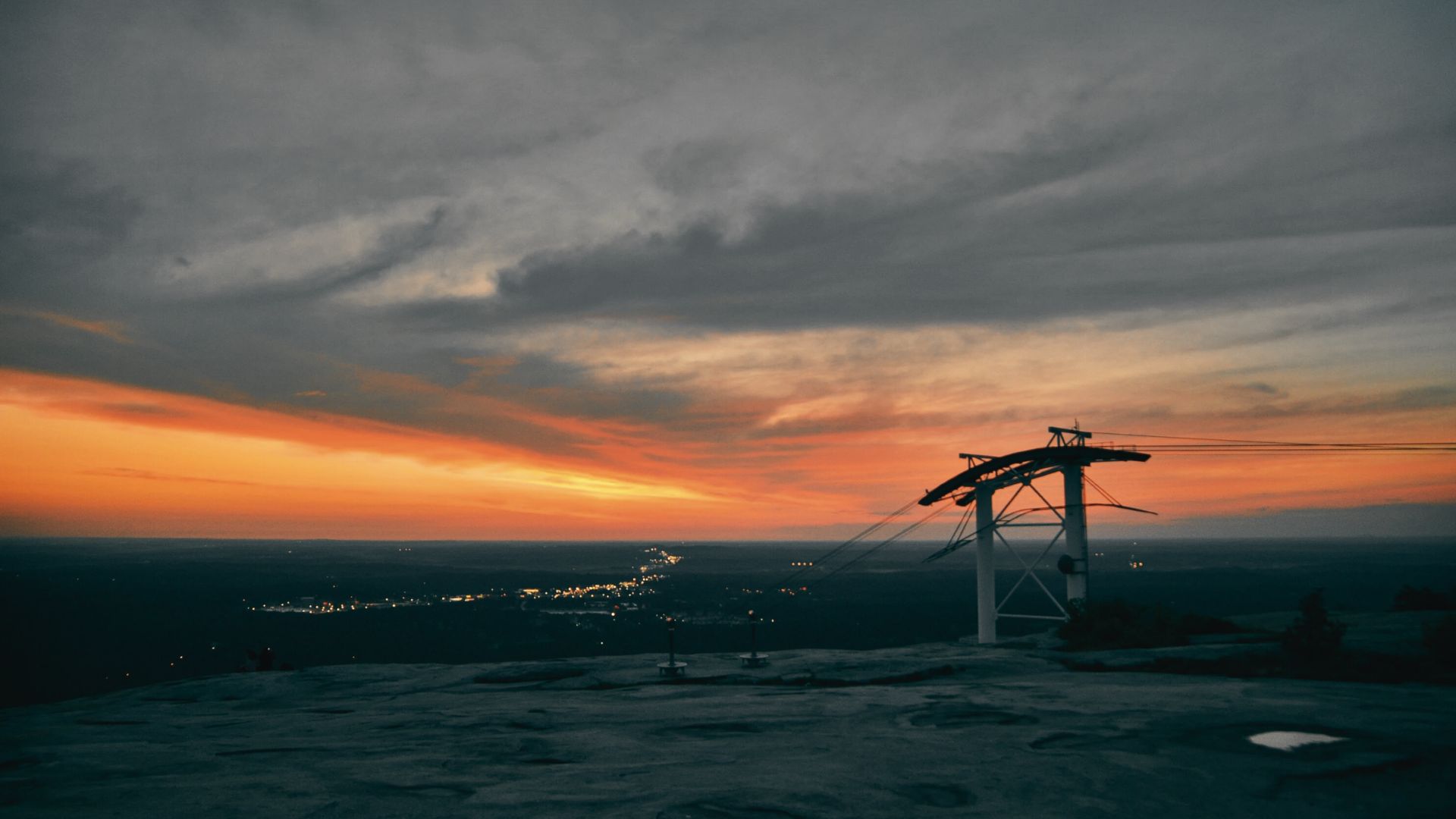 Sunset view from Stone Mountain with skyride cables silhouetted against glowing orange and gray clouds.