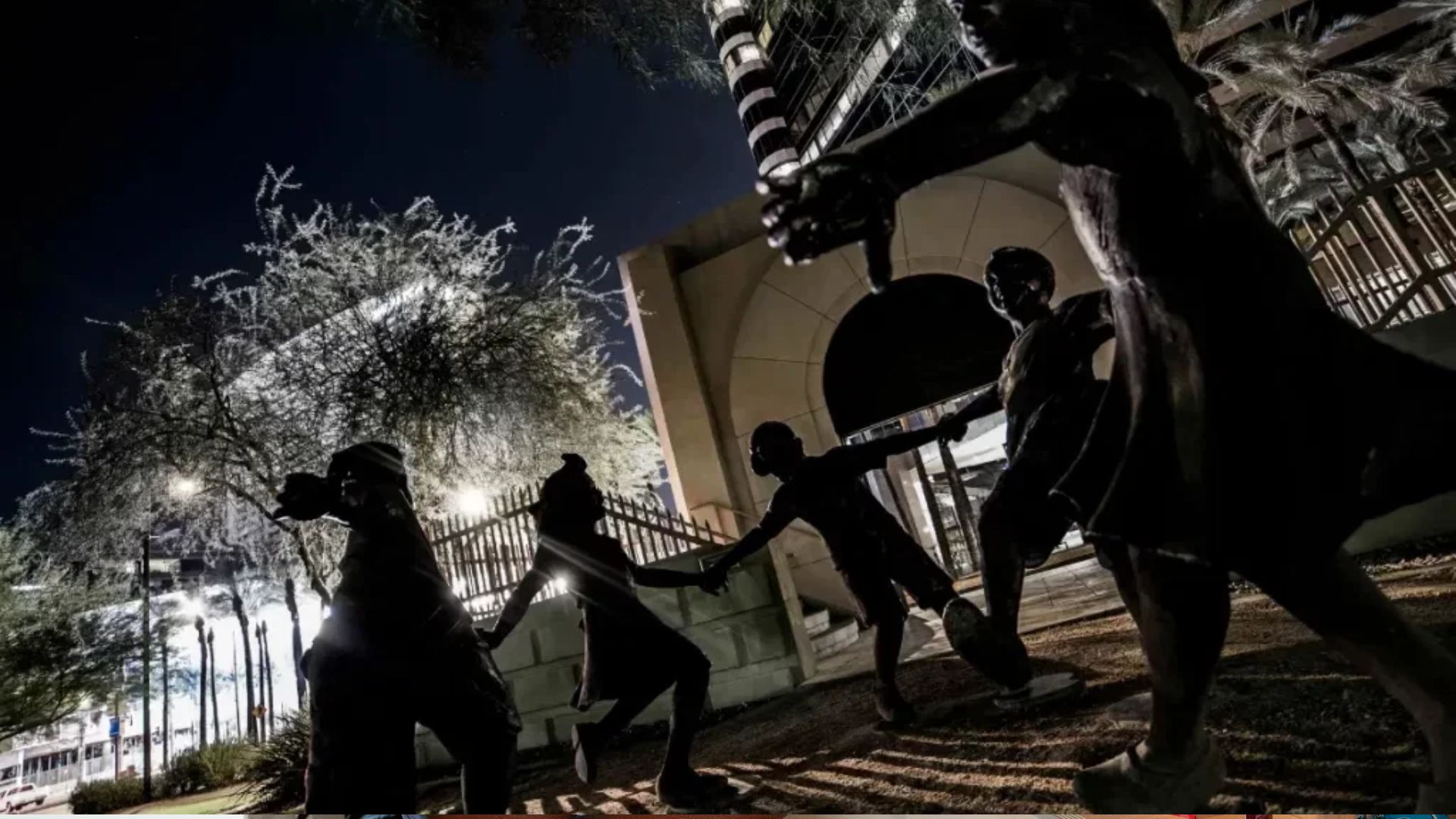 Bronze statues of five children holding hands and dancing at night, arched building entrance and illuminated trees behind.