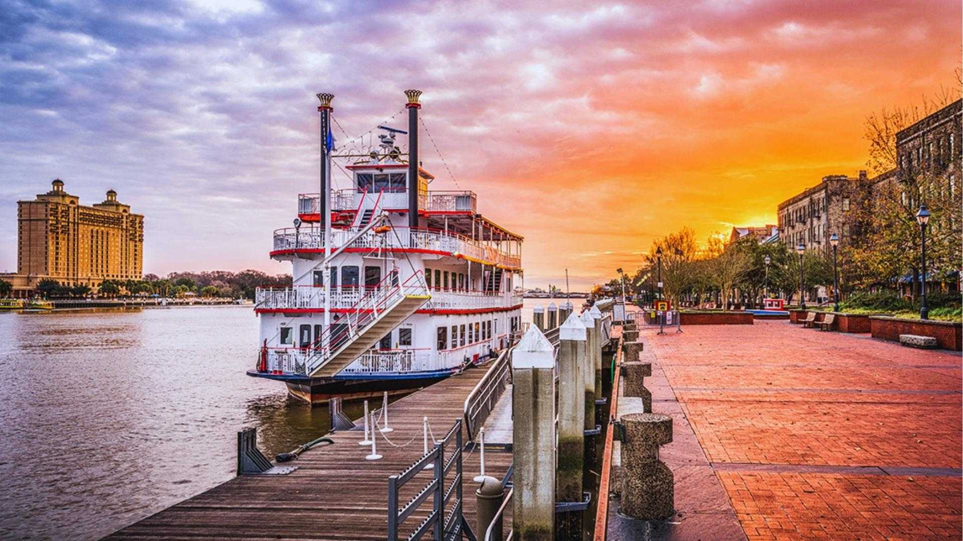 Savannah Riverboat docked at River Street at sunset with vibrant orange sky over riverfront.
