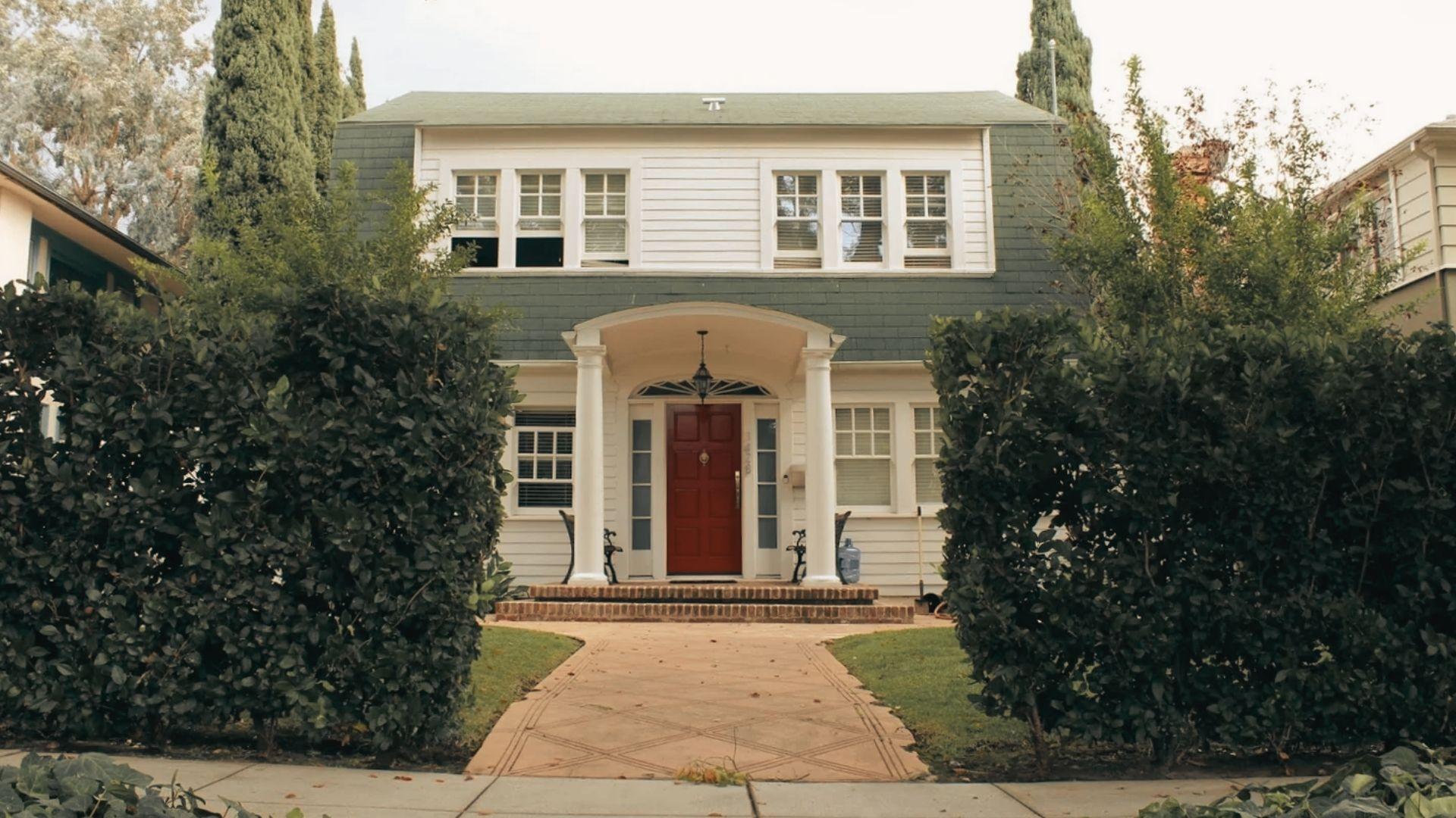 White Dutch Colonial house with green shingle roof, red front door, columns, brick walkway, hedges, tall cypress trees
