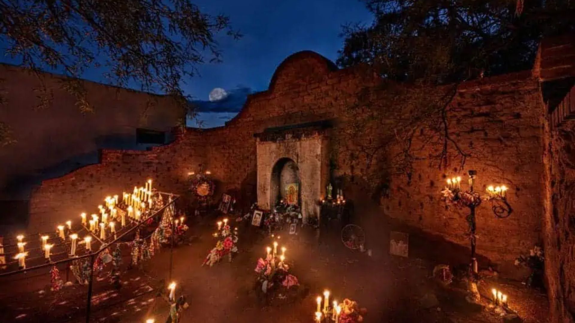 Candlelit courtyard of old stone ruins at night with many glowing candles, flowers, and full moon in dark blue sky.