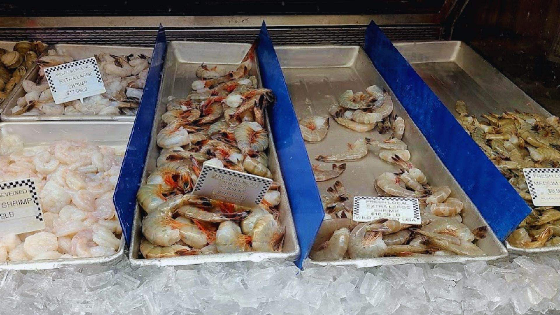 Trays of fresh shrimp on ice along with other varieties having price labels at a seafood market.