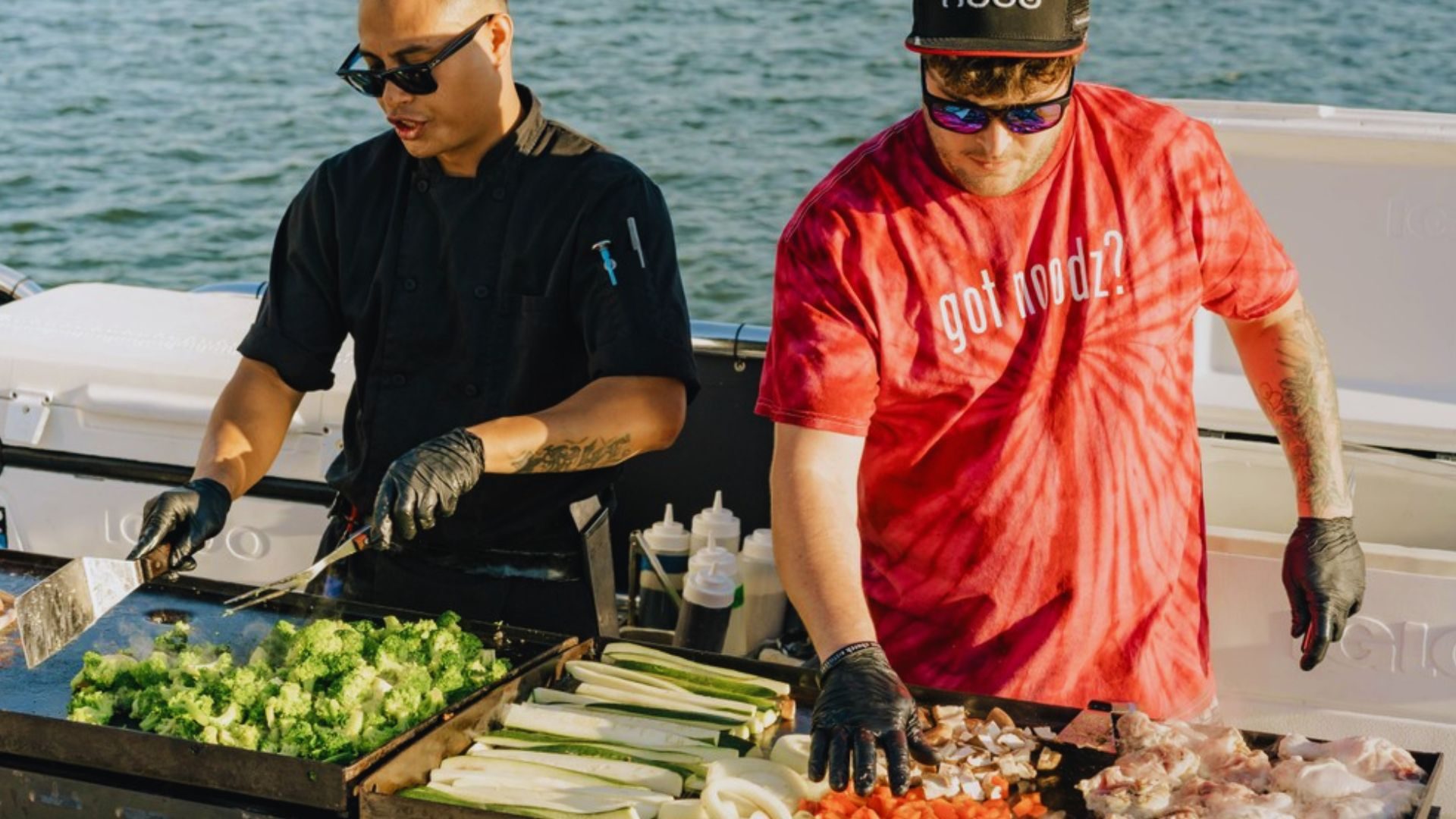 Two men cook vegetables and meat on a grill near the water at dinner cruise.