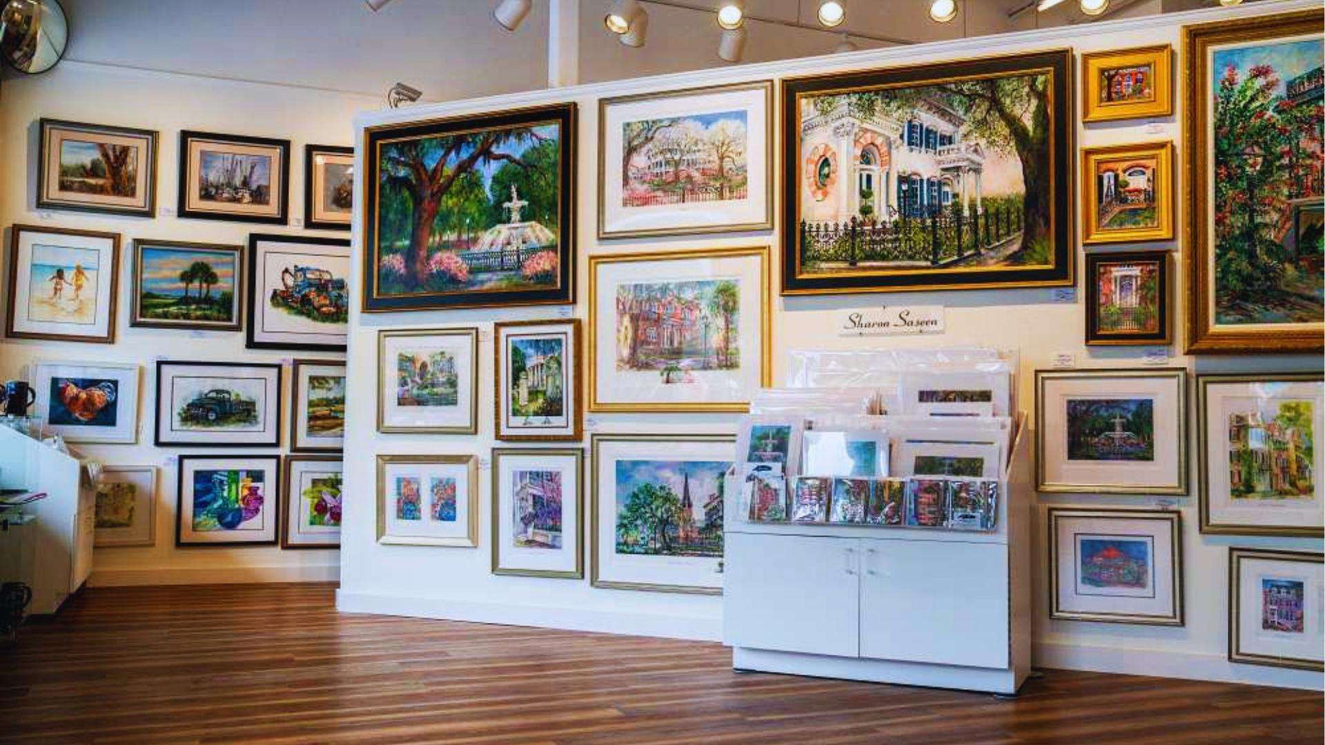 Art gallery wall filled with colorful Savannah paintings of Forsyth Park and historic homes.
