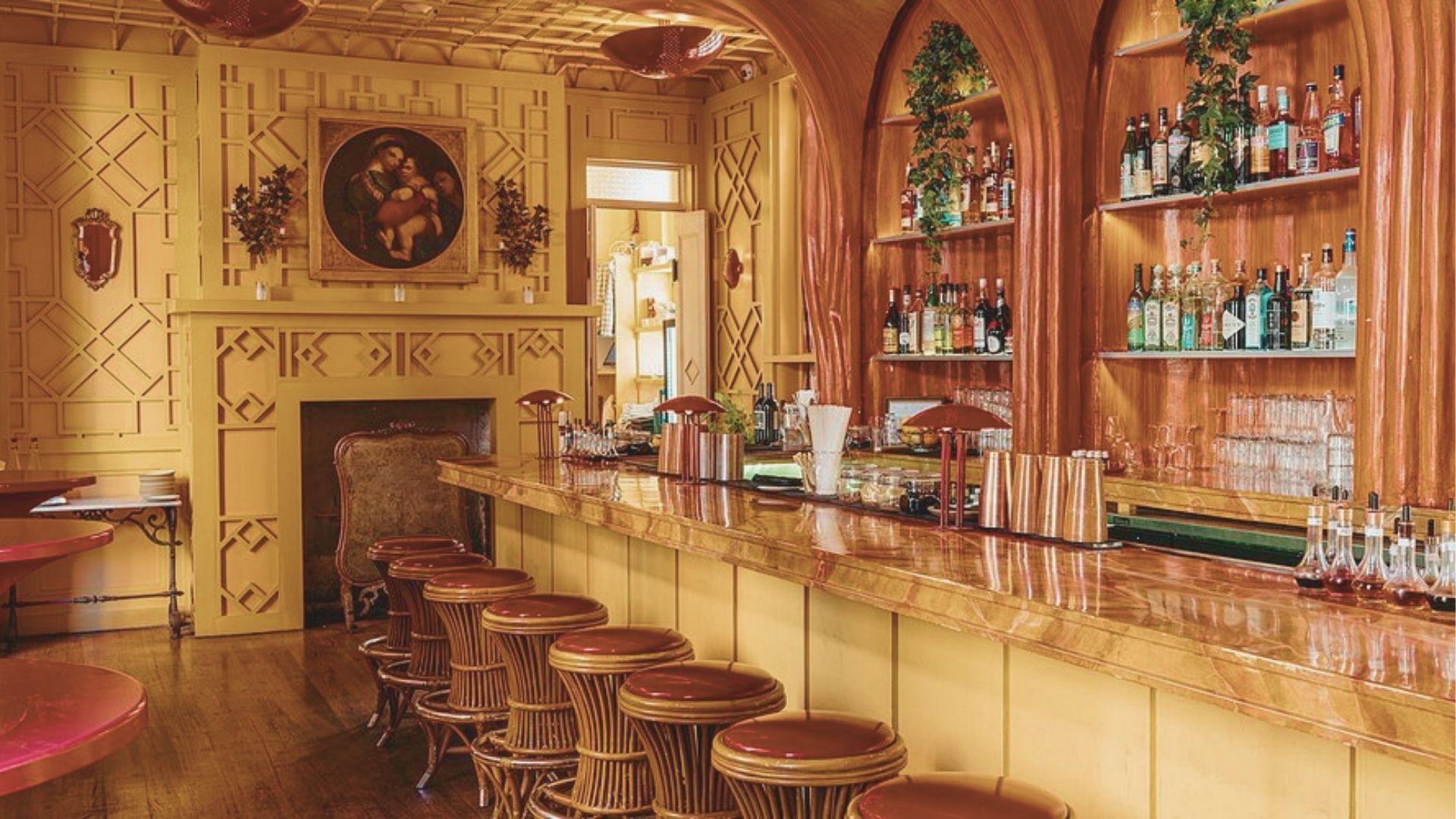 Warm, golden-toned bar interior with a marble counter, rattan stools, and ornate wood paneling.