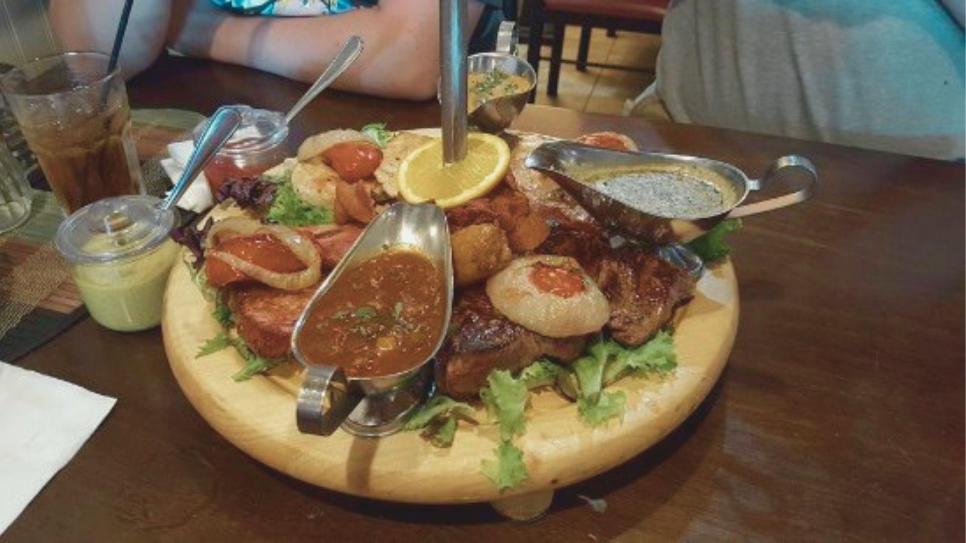 A wooden platter loaded with German meats, roasted vegetables, and gravy boats served at a restaurant table.