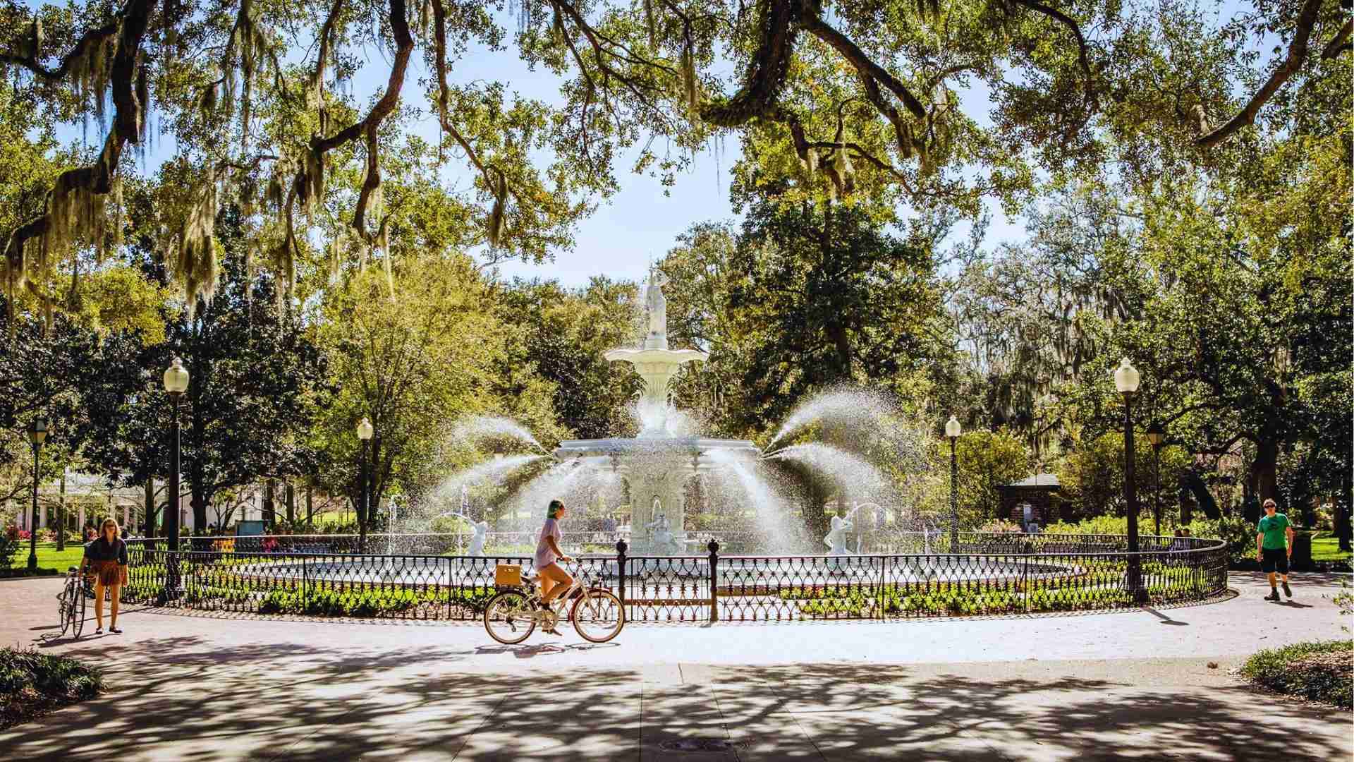 Forsyth Park fountain sparkling in sunlight with cyclists riding past under Spanish moss oaks.