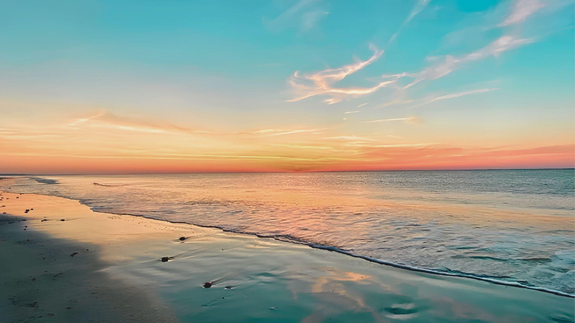 Sunset beach with calm waves, wet sand reflecting orange and turquoise sky.