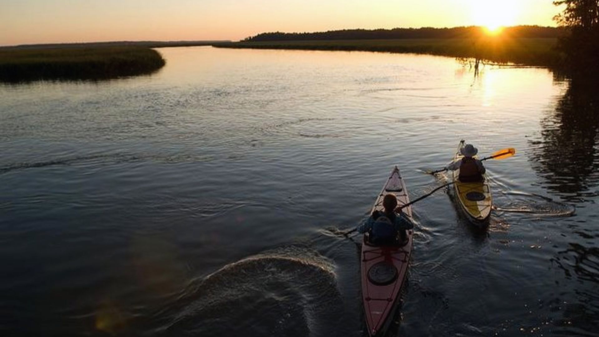 Two people kayak on calm water through a marsh at sunset.