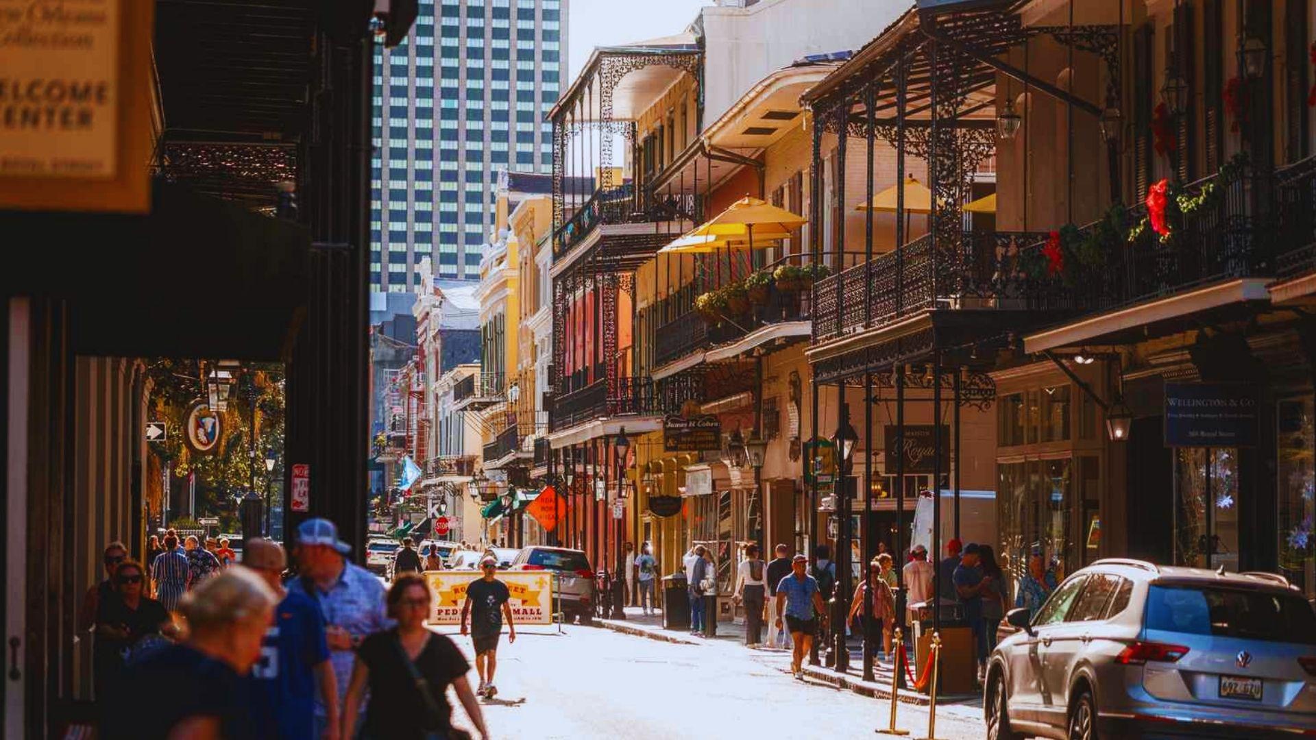 Sunlit Royal Street with ornate iron balconies, shops, pedestrians, parked cars, modern skyscraper visible, French Quarter