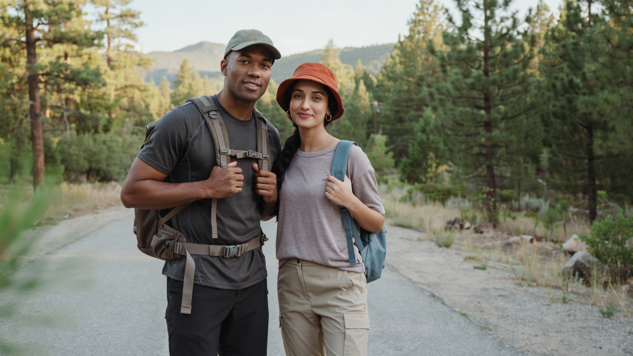 Two hikers with backpacks stand on a forest trail surrounded by trees and distant mountains.