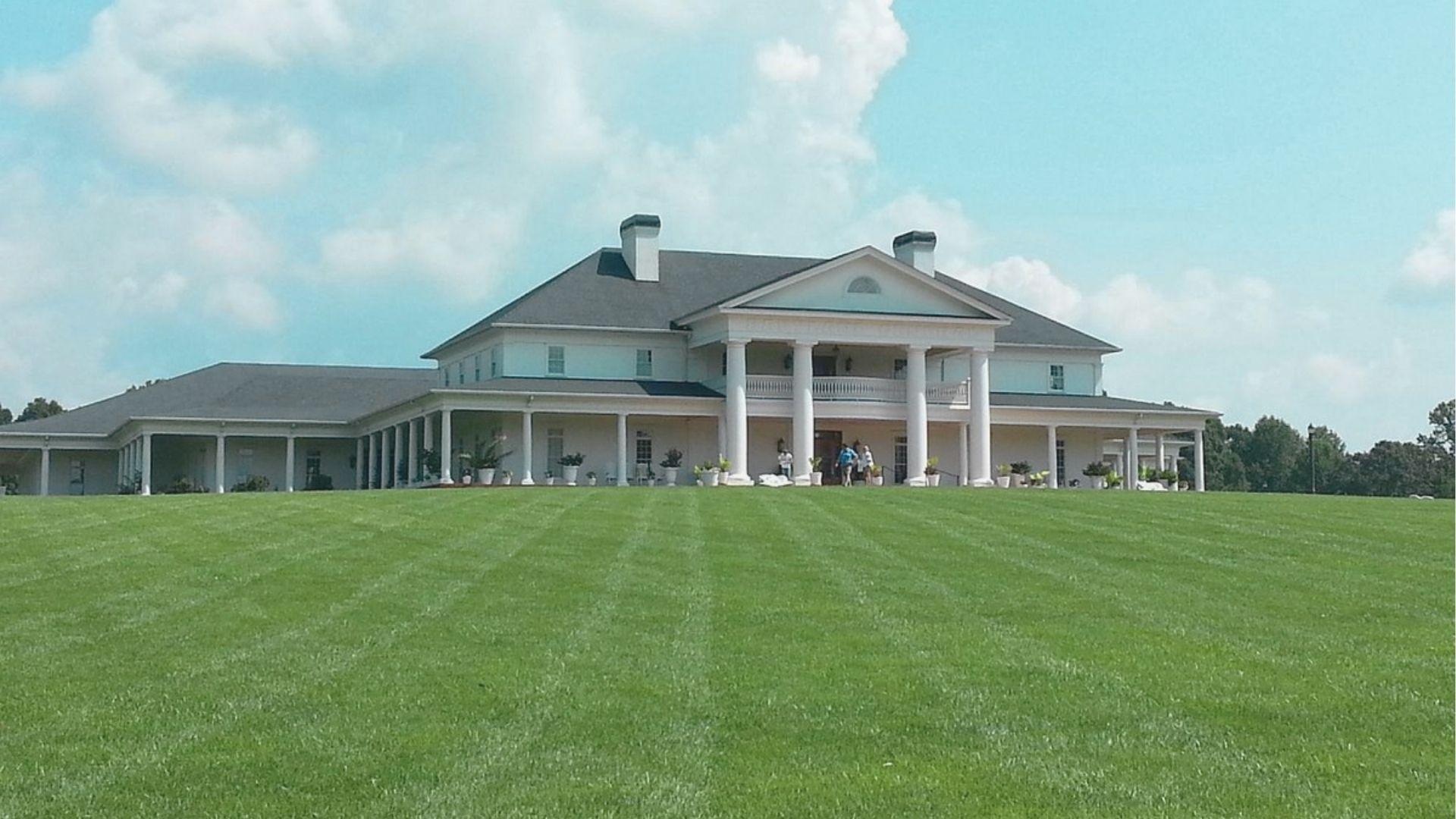 A grand white mansion with tall columns sits on a manicured green lawn under a blue sky.