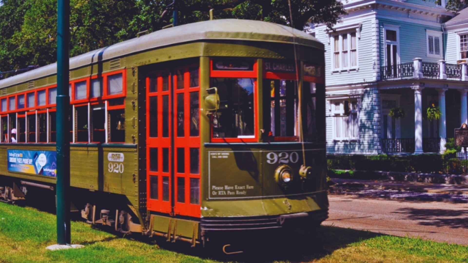 Green and red St. Charles streetcar 920 passes blue Victorian mansion with columns, trees, sunny day