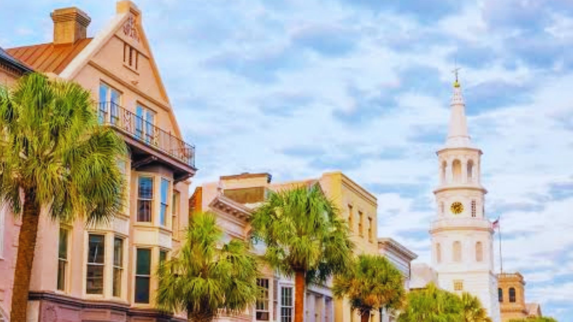 Historic Charleston street with palm trees, colorful buildings, church steeple, and cloudy daytime sky.
