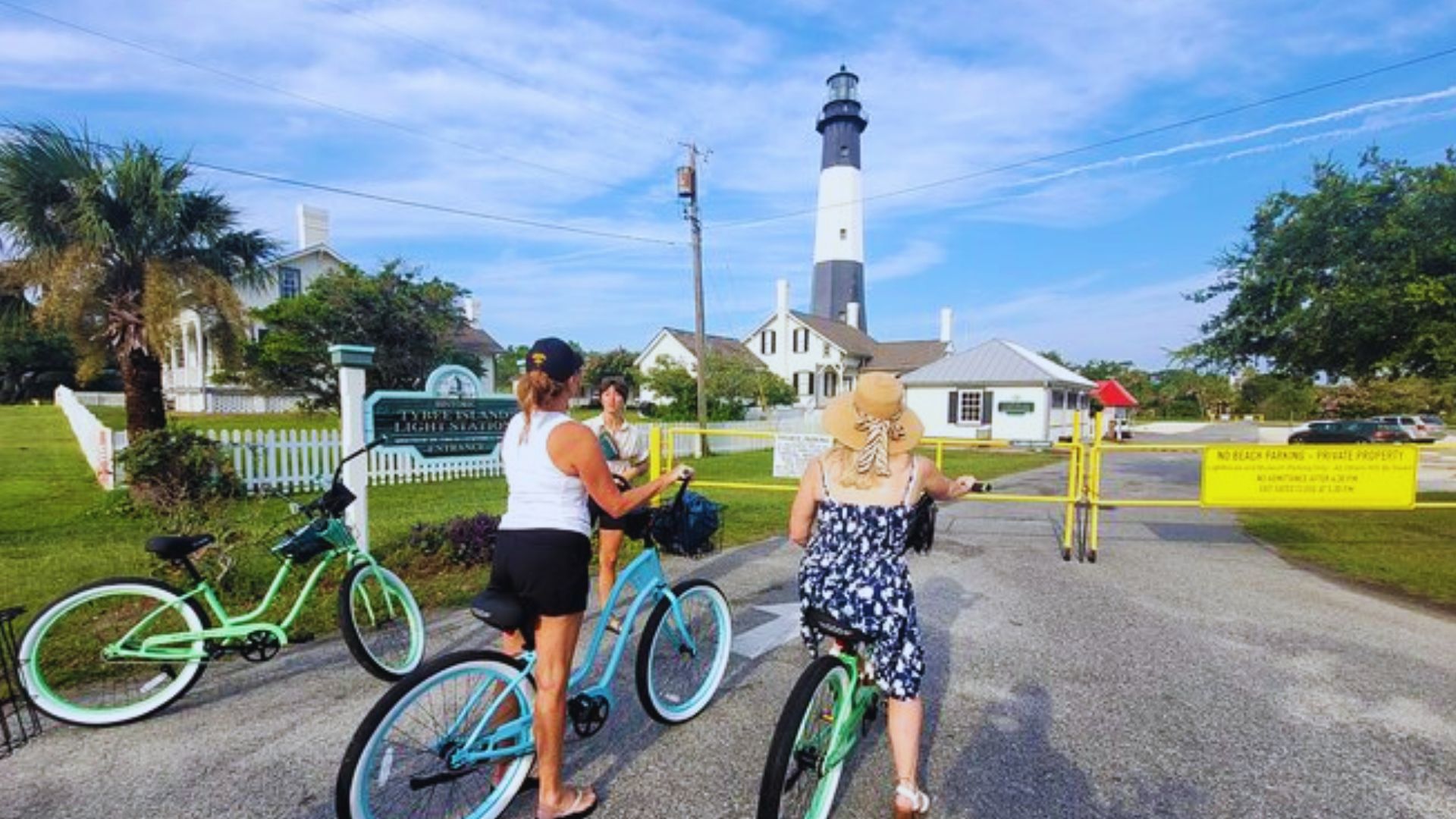 People ride bicycles near Tybee Island Light Station with historic buildings and palm trees nearby.