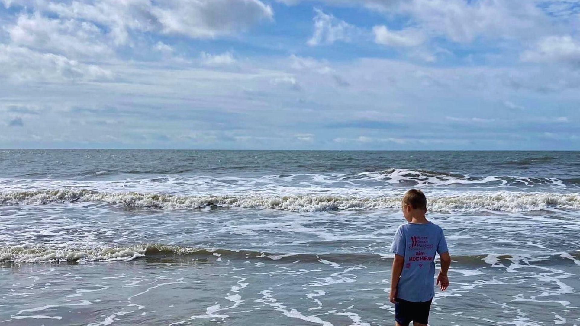 A boy in a shirt watches ocean waves roll in under the cloudy sky at Driessen Beach Park.