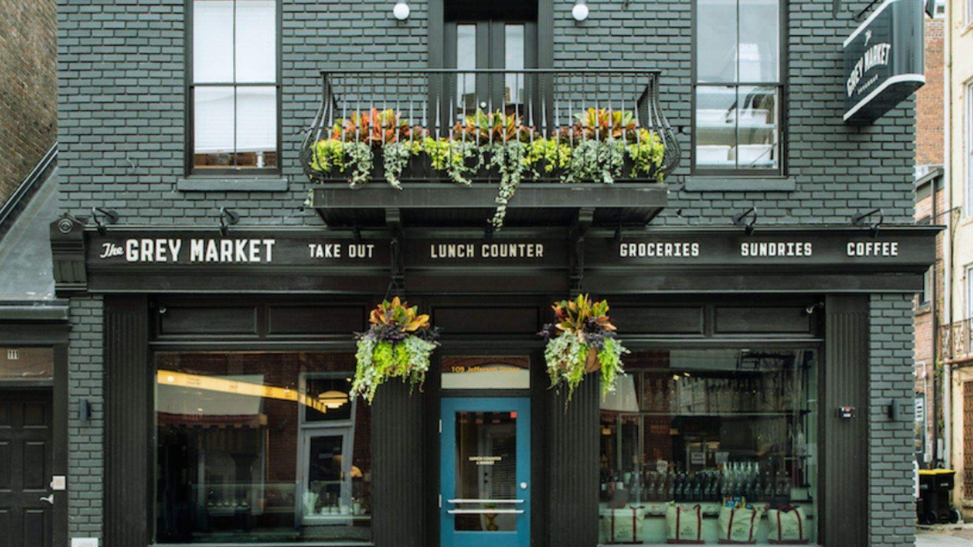Dark gray brick storefront named The Grey Market with blue door, flower baskets, and balcony plants.