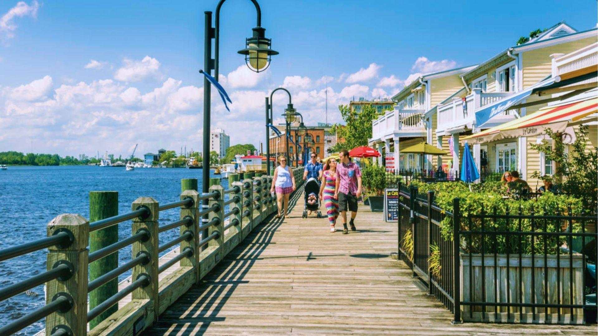 Riverfront boardwalk in Wilmington with families strolling past shops cafes and vintage street lamps overhead.
