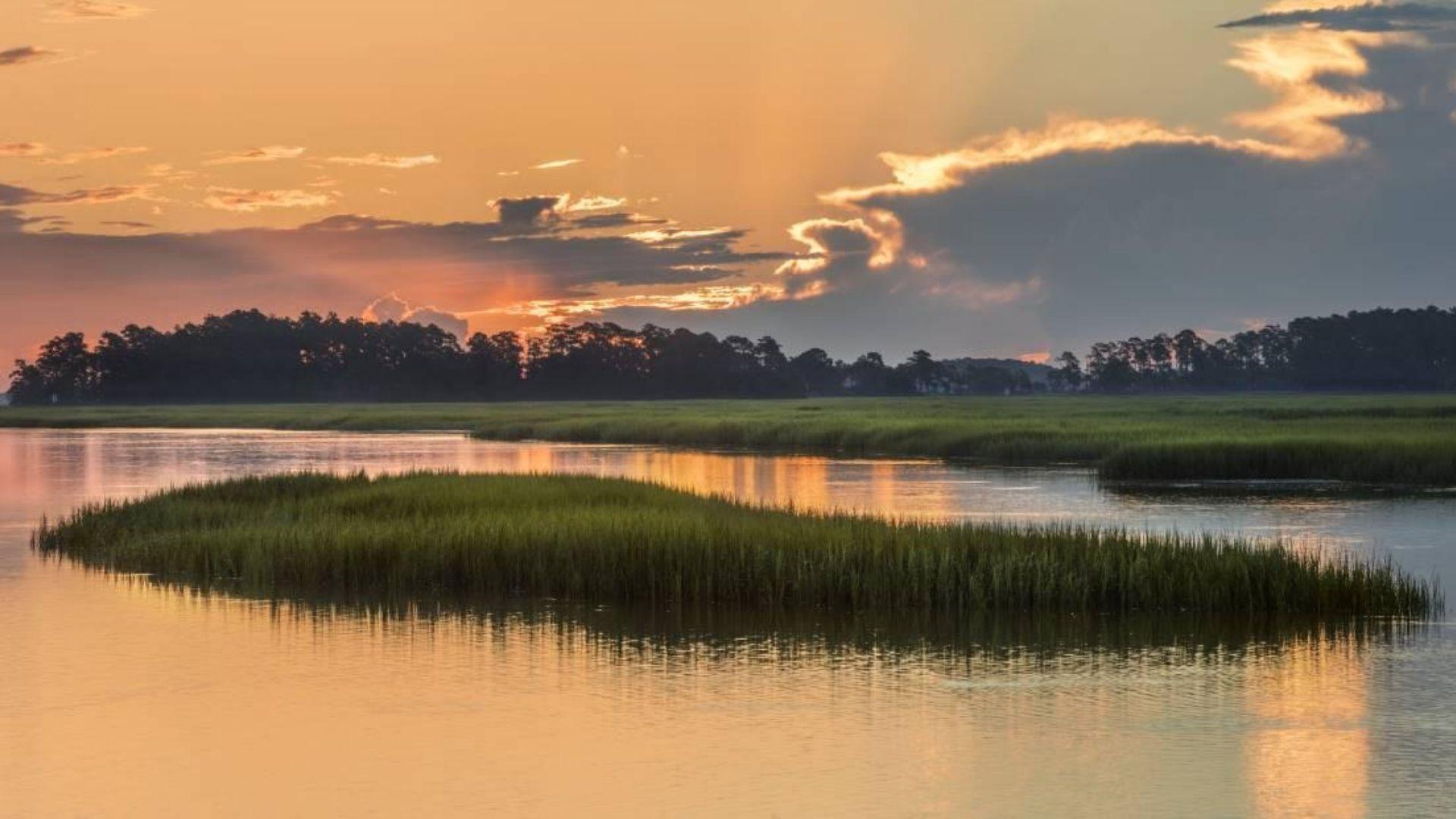 Golden sunset over marsh waterways, grassy islands reflecting orange sky and dramatic clouds